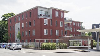 Mehrfamilienhaus mit roter Fassade, drei Stockwerke, Balkone und Fenster. Im Vordergrund stehen geparkte Autos und ein überdachter Fahrradabstellplatz.