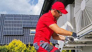 Foto, im Vordergrund ein Mann auf einer Leiter, der Wärmepumpen prüft, im Hintergrund ein Haus mit einer Solaranlage auf dem Dach.