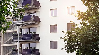 Foto, Fassade eines neuen, mehrstöckigen Wohngebäudes mit Balkonen und daran montierten Solarmodulen.