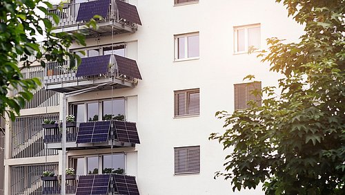 Foto, Fassade eines neuen, mehrstöckigen Wohngebäudes mit Balkonen und daran montierten Solarmodulen.