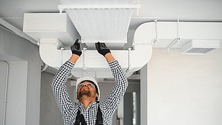 Foto, Ein Arbeiter im karierten Hemd und mit Handschuhen arbeitet an einer Klimaanlage an der Decke eines modernen, unfertigen Raums.