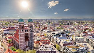 Foto, Luftaufnahme von München mit den Zwiebeltürmen der Frauenkirche im Vordergrund und der Alpenkette am Horizont