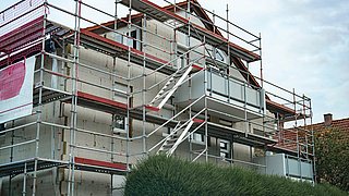 Foto, Mehrstöckiges Wohnhaus mit Gerüst und Baustellenaufzug, davor dichter grüner Busch und mobiler Toilettencontainer