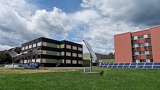 Zwei Gebäude auf einer Wiese: links ein dreigeschossiges, schwarz-grünes Gebäude mit einem Flugzeug auf dem Dach, rechts ein rotes, dreigeschossiges Gebäude mit Balkonen. Im Vordergrund stehen Solarpanels.