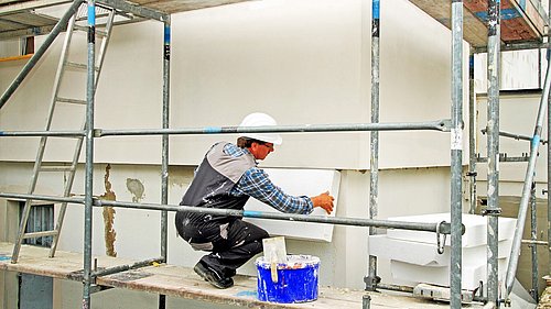 Foto, ein Bauarbeiter kniet auf einem Baugerüst und montiert Dämmplatten an einem Gebäude.