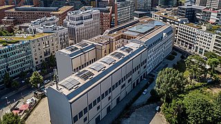 Foto, Luftaufnahme vom Potsdamer Platz in Berlin mit einem langen, rechteckigen Gebäude, der Kältezentrale der Berliner Energie und Wärme GmbH im Vordergrund, umgeben von modernen Bürogebäuden. Im Hintergrund sind die beiden Hochhäuser der Deutschen Bahn und des Kollhoff Towers, das Ritz Hotel sowie weitere Gebäude sichtbar.