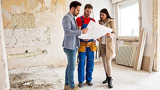 Foto, eine Frau und ein Mann schauen in einem unsanierten Haus gemeinsam mit einem Handwerker auf ein Dokument.