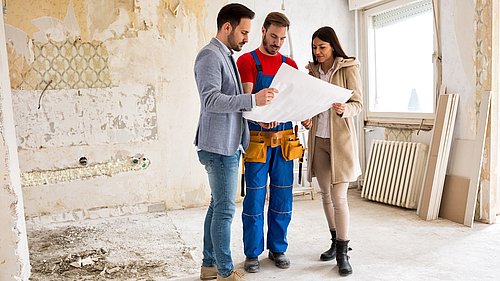 Foto, eine Frau und ein Mann schauen in einem unsanierten Haus gemeinsam mit einem Handwerker auf ein Dokument.