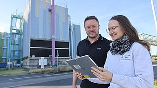 Foto, eine Frau und ein Mann stehen draußen vor einen Industrieanlage und schauen auf einen Tablet-Computer